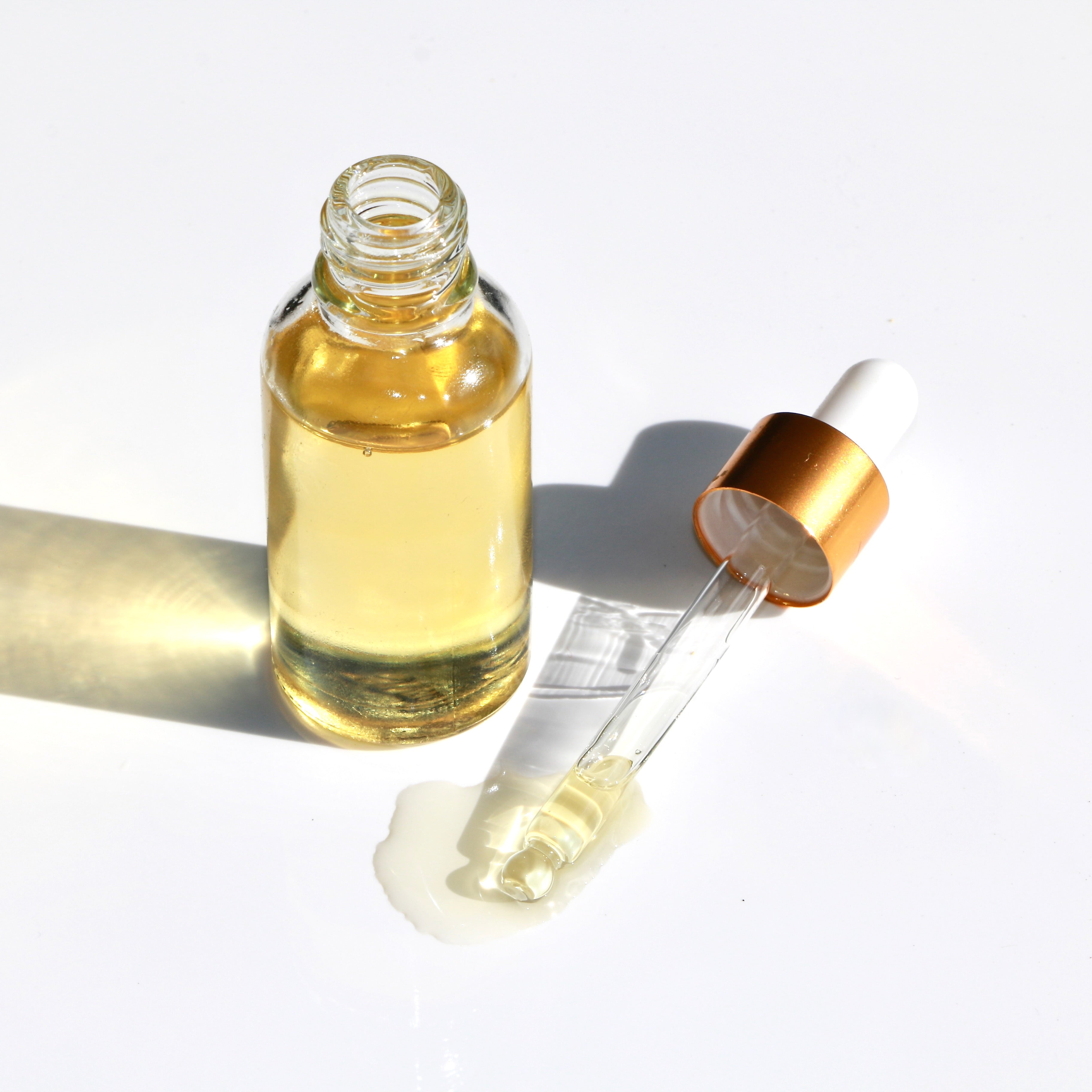 Dropper bottle of oil with dropper placed next to base, surrounded by pooled oil on a sleek surface.