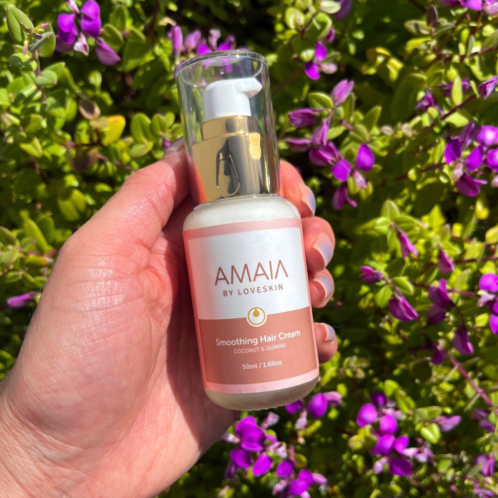 Hand holding a bottle of Amaia by Loveskin Smoothing Hair Cream against a floral background