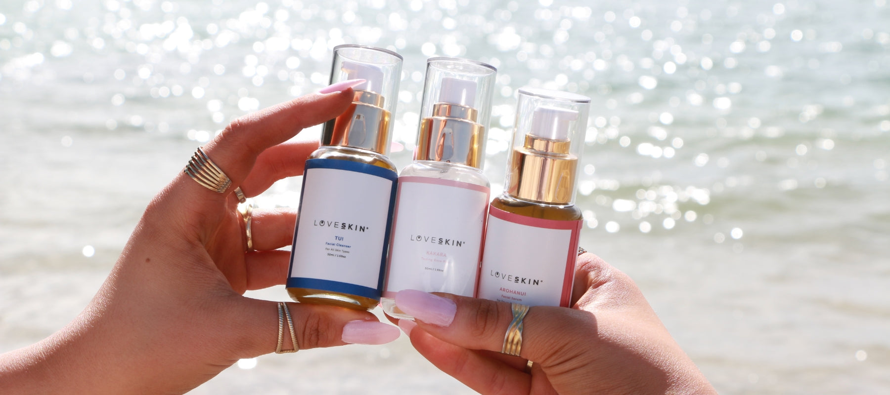 Close-up of tanned hands holding 50ml LoveSkin Header 3-Step Skincare Ritual bottle with shimmering sea background.